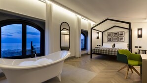 A modern bedroom with a bathtub, a bed with a checkered bedspread, a green chair, and a view of the city through large windows. - Maison Bon Bon (Sorrento)