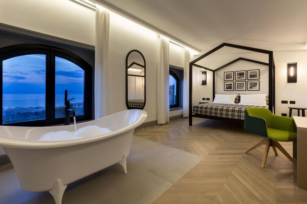 A modern bedroom with a bathtub, a bed with a checkered bedspread, a green chair, and a view of the city through large windows.