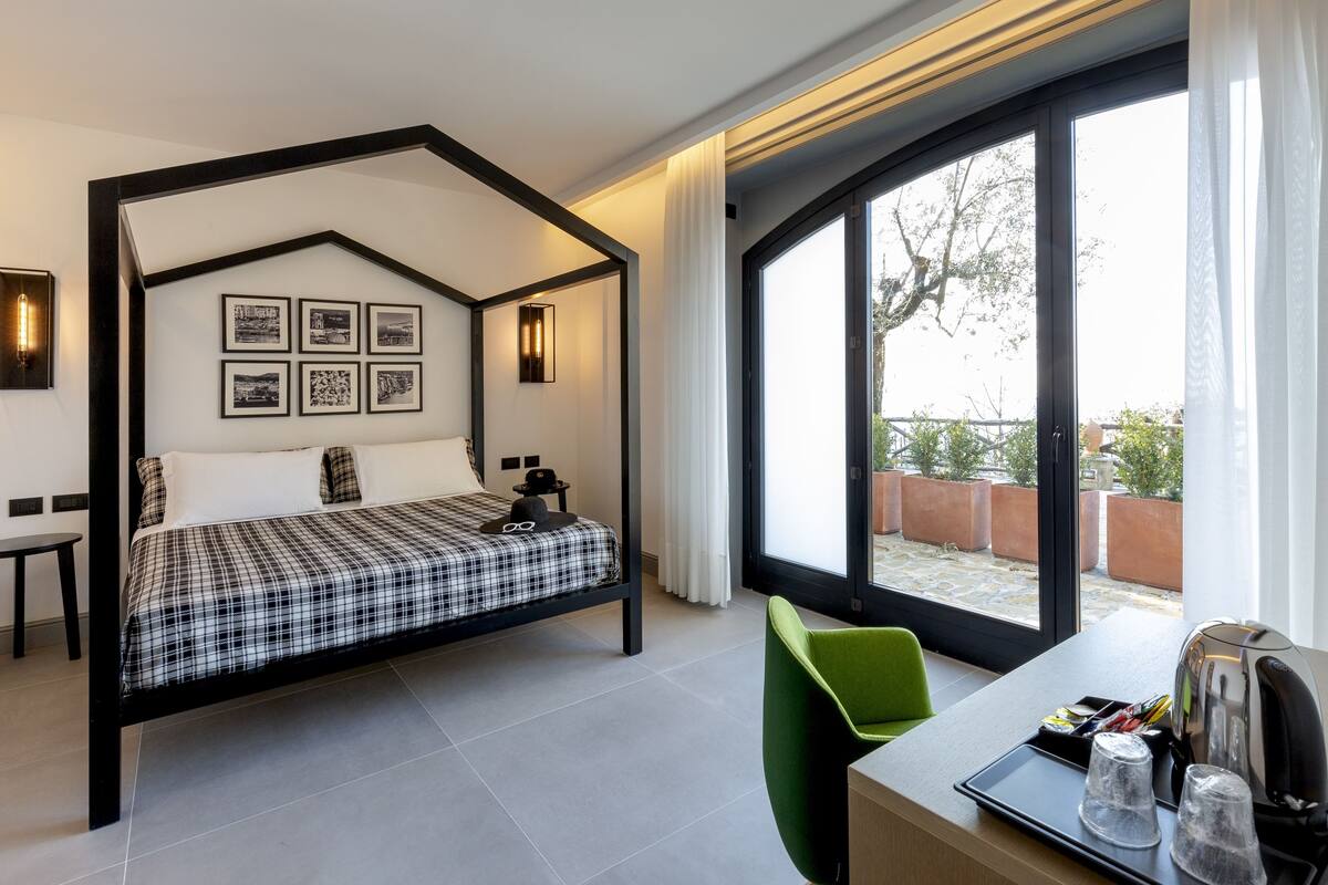A modern hotel room with a four-poster bed, a green chair, a desk with a kettle, and a large window offering a view of the outdoors.