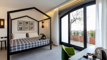 Ein modernes Hotelzimmer mit Himmelbett, einem grünen Sessel, einem Schreibtisch mit Wasserkocher und einem großen Fenster mit Blick ins Freie.