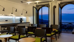 A modern dining area with round tables and chairs, a bar with a white countertop, and large arched windows offering a view of the sea. - Maison Bon Bon (Sorrento)