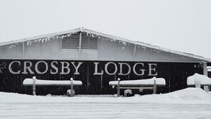 A large, industrial building labeled "Crosby Lodge" with a flat roof and ventilation grilles. Snow is piled in front of the building, and there are metal pipes and a fence visible. - Crosby Lodge (Crosby)