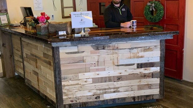 A reception desk with a wooden panel design, a sign for assistance, and a wreath on the wall. - Crosby Lodge (Crosby)
