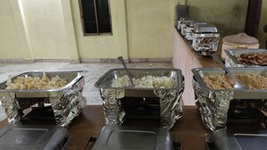 A buffet setup with multiple food dispensers covered in aluminum foil, a tiled floor, and a door in the background. - Aroma Desert Safari Camp (Jaisalmer)