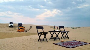 A desert scene with two vehicles, a camel, and a table with chairs and a blanket. - Aroma Desert Safari Camp (Jaisalmer)