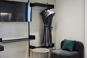 Una habitación moderna con una televisión de pantalla plana montada en un estante negro, una bata gris colgada de un perchero, una pequeña mesa redonda blanca y un sofá gris oscuro con un cojín turquesa.
