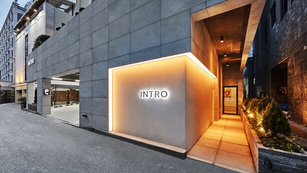 Ingresso moderno di un edificio con un cartello 'INTRO' ben visibile, tettoia e area verde con piante.