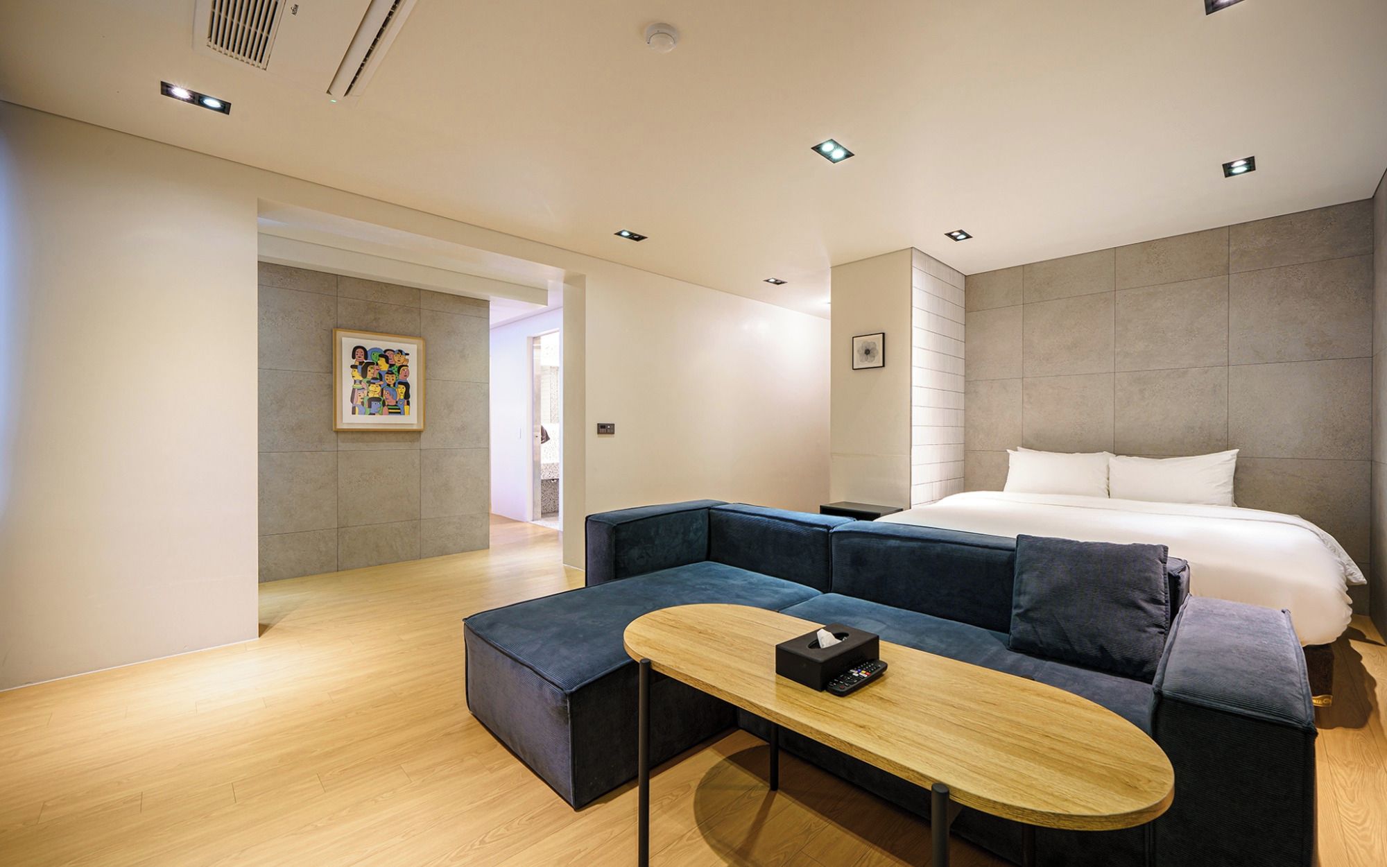 Una habitación de hotel moderna con una cama grande, un sofá azul oscuro, una mesa de centro de madera y un cuadro en la pared.