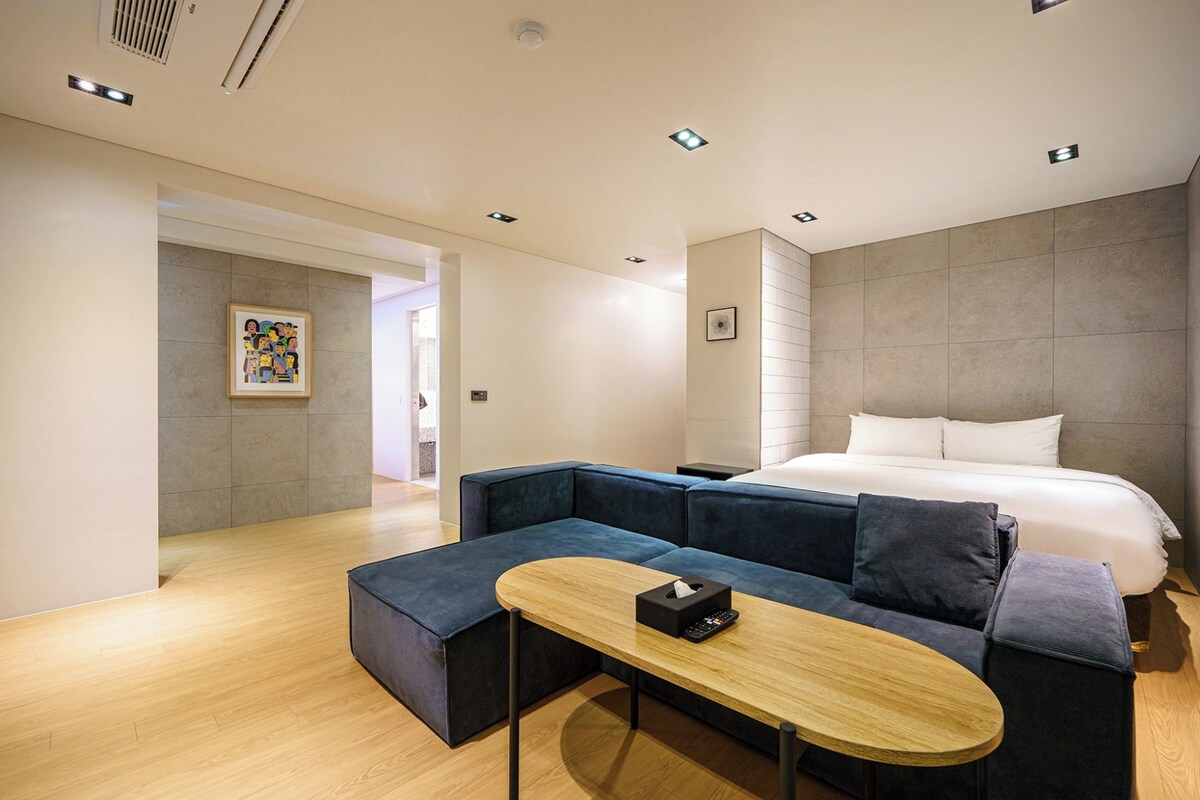 Una habitación de hotel moderna con una cama grande, un sofá azul oscuro, una mesa de centro de madera y un cuadro en la pared.