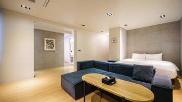 Una habitación de hotel moderna con una cama grande, un sofá azul oscuro, una mesa de centro de madera y un cuadro en la pared.