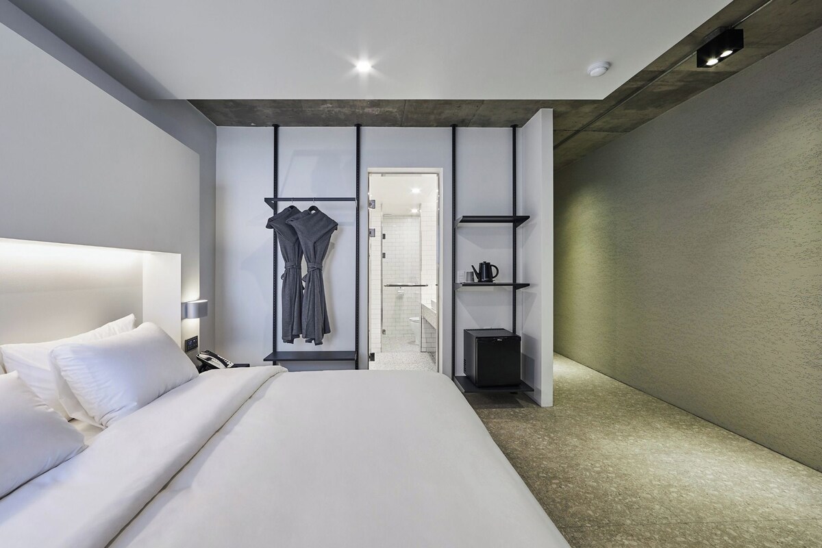 Una habitación de hotel moderna con una cama grande, un perchero para bata, un estante pequeño con artículos de aseo y vistas al baño.
