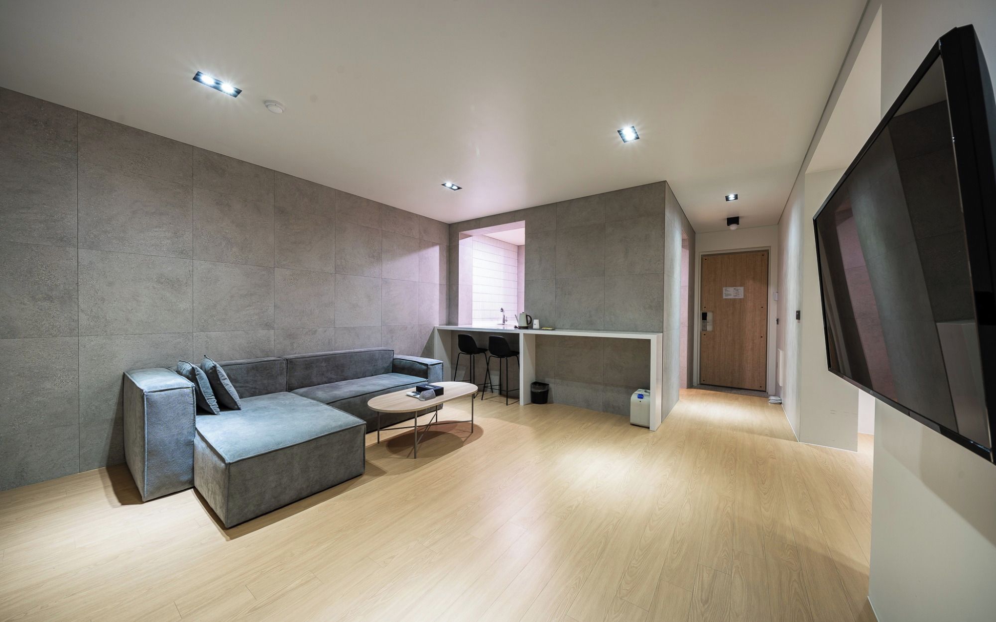 Ein modernes Wohnzimmer mit einem grauen Ecksofa, einem kleinen runden Tisch und einer Küchenzeile inklusive Arbeitsplatte und Barhocker.