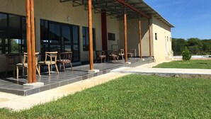A covered outdoor seating area with chairs and tables, a building with large windows, and a grassy lawn. - Shumba Lodge (Tutume)