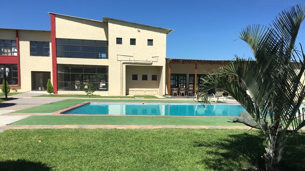A modern building with a swimming pool, green lawn, and palm trees. - Shumba Lodge (Tutume)