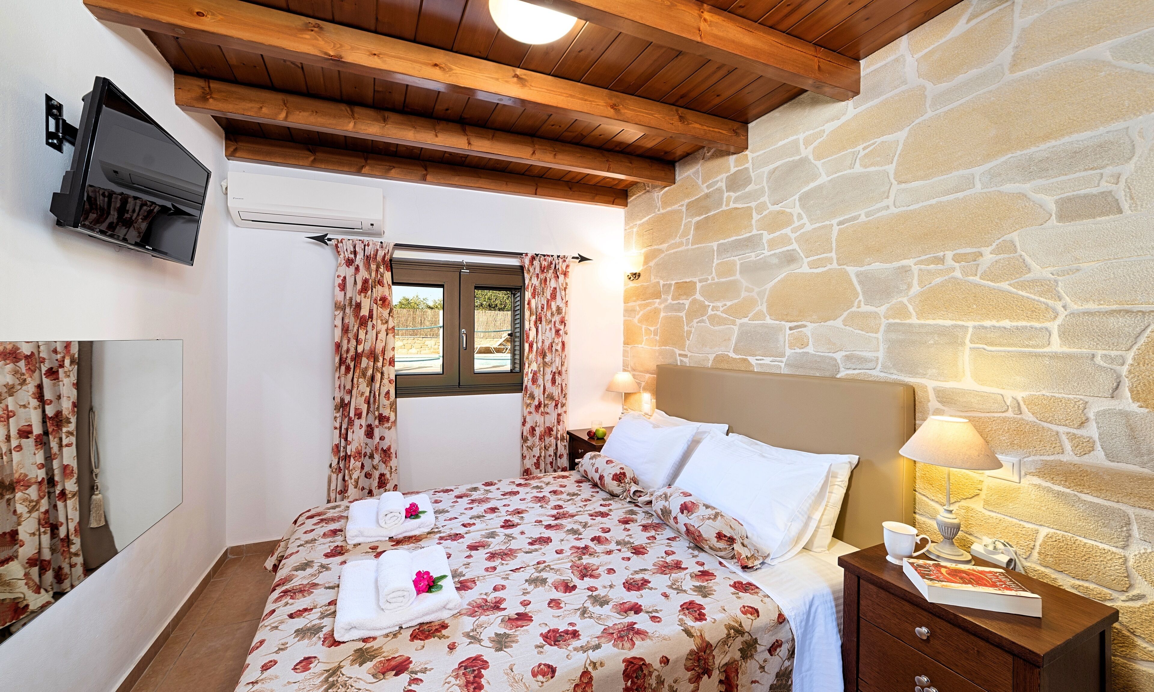 Een slaapkamer met een stenen muur, houten balken in het plafond, een bed met bloemenbedrukking, een aan de muur bevestigde televisie en een raam met gordijnen.