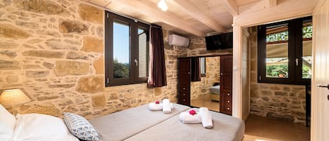 A bedroom with stone walls, wooden ceiling beams, a bed with pillows, and a window with shutters.
