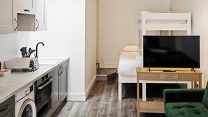 Ein kompaktes Hotelzimmer mit Küchenzeile, Waschmaschine, Etagenbett, Fernseher und einem grünen Sessel.