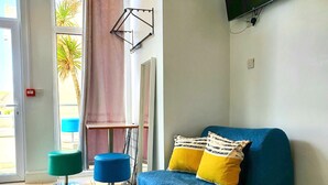 Ein modernes Wohnzimmer mit einem blauen Sofa, zwei Hockern, einem an der Wand montierten Fernseher und Blick auf Palmen durch das Fenster.