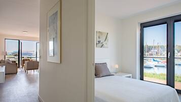 A modern hotel room with a large bed, a desk, and a chair. There is a view of boats through the window.