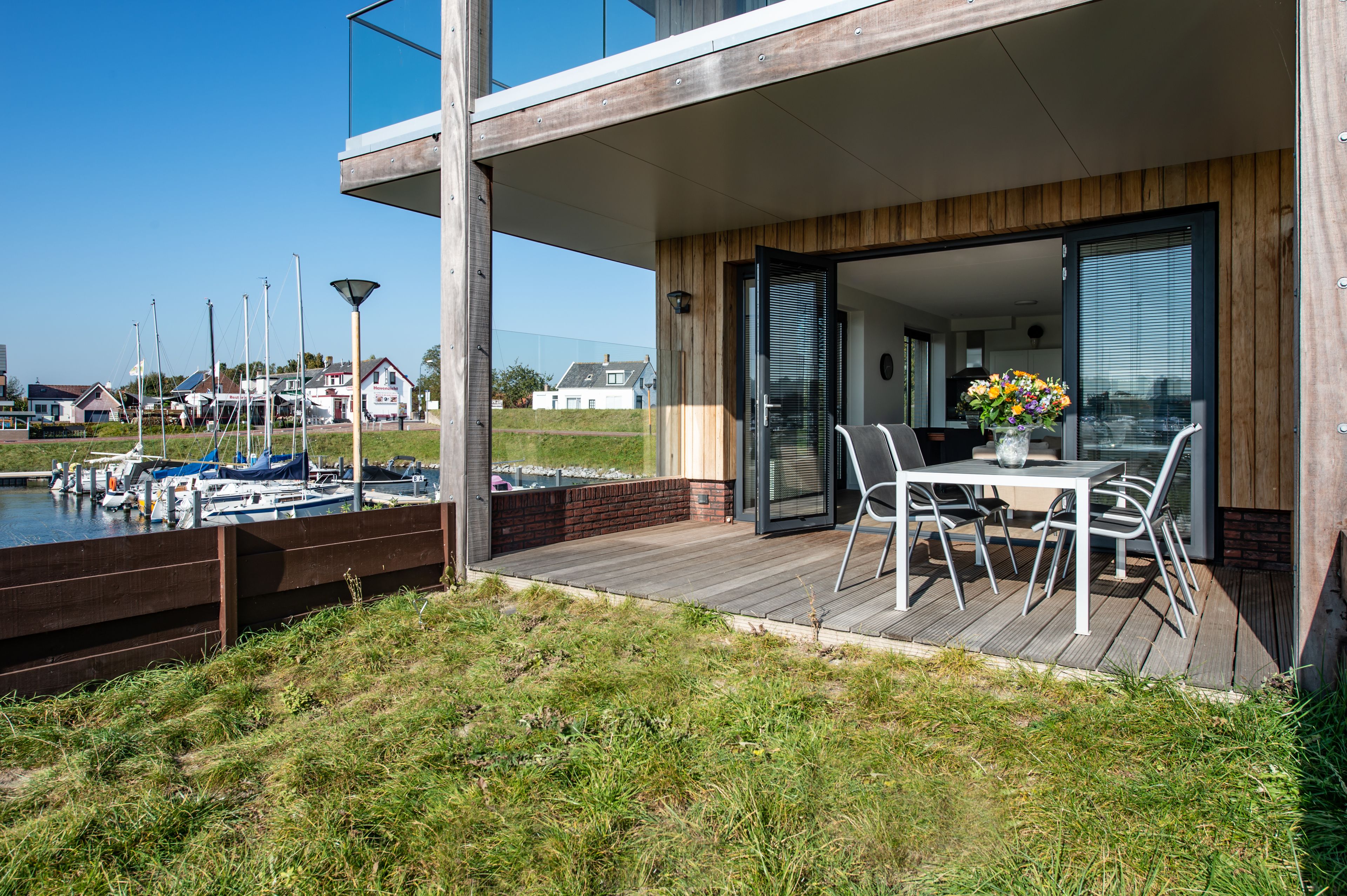 A modern outdoor patio with a table and chairs, a wooden deck, and a view of a marina with boats.