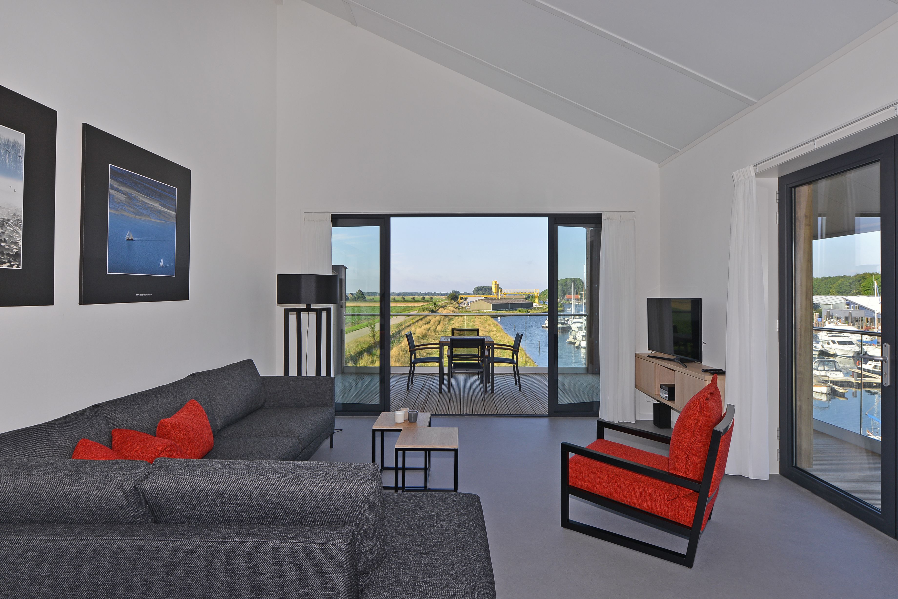 A modern living room with a grey sofa, red chair, and a small table. There is a large window with a view of a marina and a distant landscape.