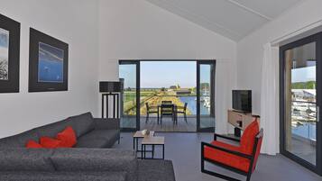 A modern living room with a grey sofa, red chair, and a small table. There is a large window with a view of a marina and a distant landscape.