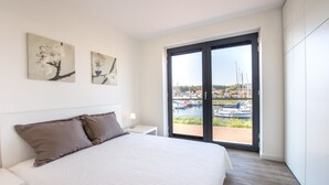 Un dormitorio con una cama grande, una mesita de noche, una lámpara y dos cuadros colgados en la pared. Tiene una puerta de cristal corredera con vistas a un puerto deportivo.