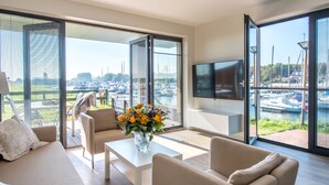 Un salón moderno con un ventanal que da a un puerto deportivo, un televisor de pantalla plana montado en la pared y un florero sobre una mesa de centro.