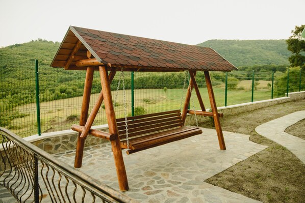 Un columpio de madera con techo de metal, situado en una zona pavimentada con piedra, una valla verde y un paisaje montañoso al fondo.