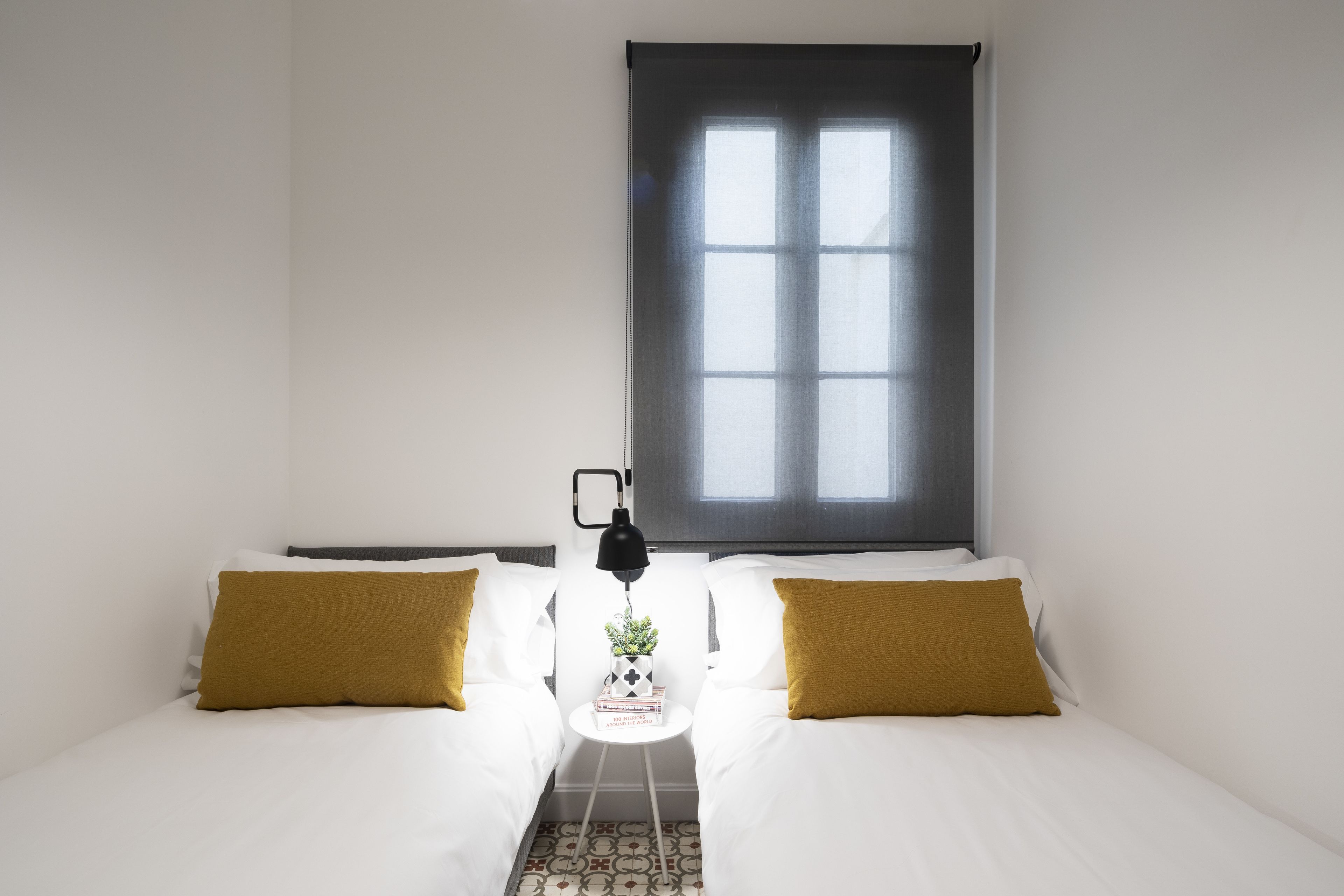 A bedroom with two beds, a window with blinds, a small table with a lamp, and a decorative item on the table.