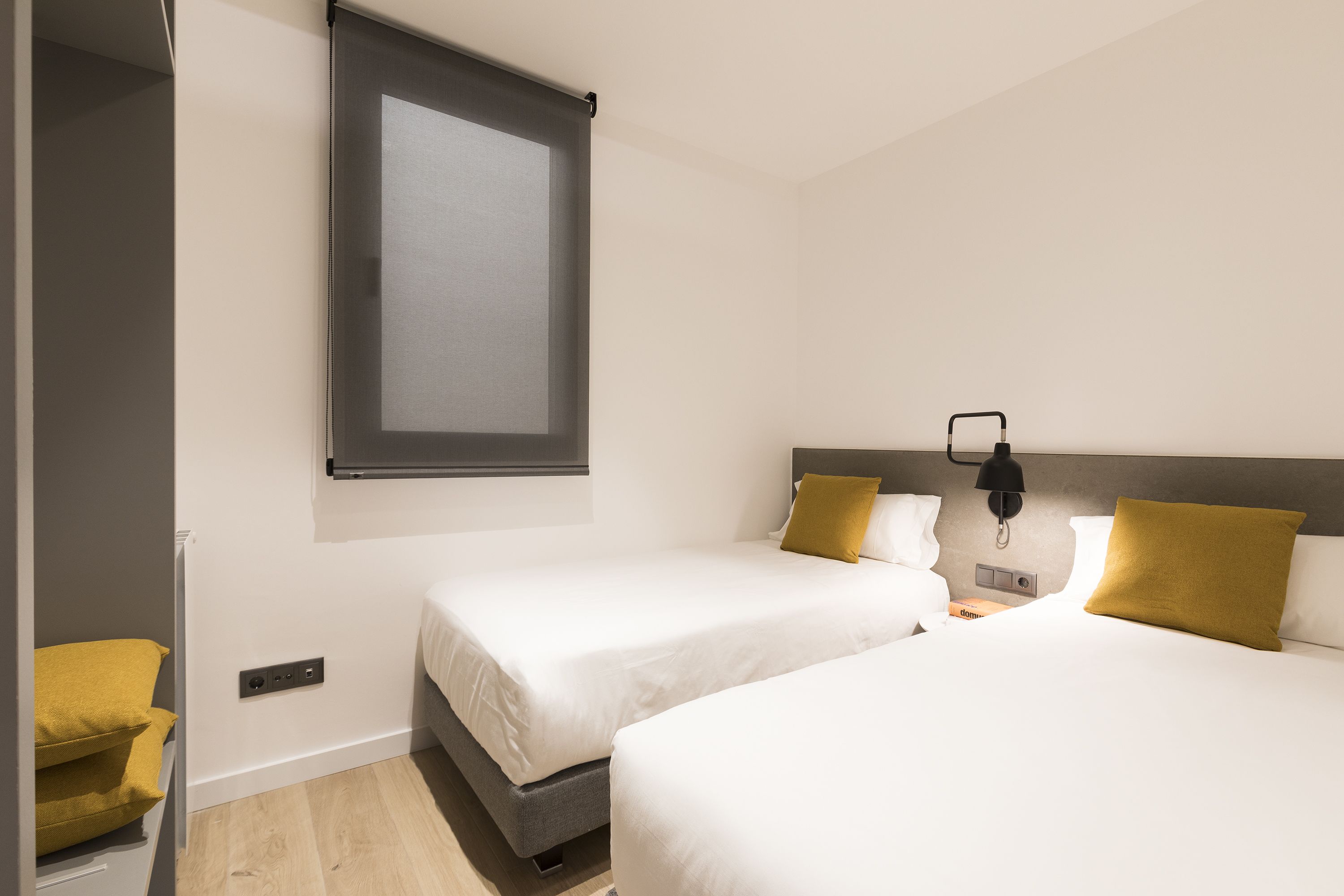 A hotel room with two beds, a window, a bedside table with a lamp, and a power outlet.