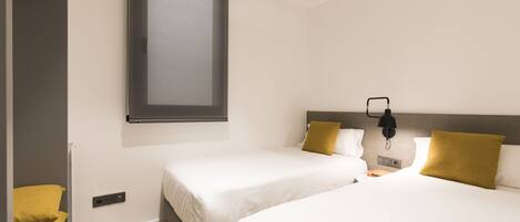 A hotel room with two beds, a window, a bedside table with a lamp, and a power outlet.
