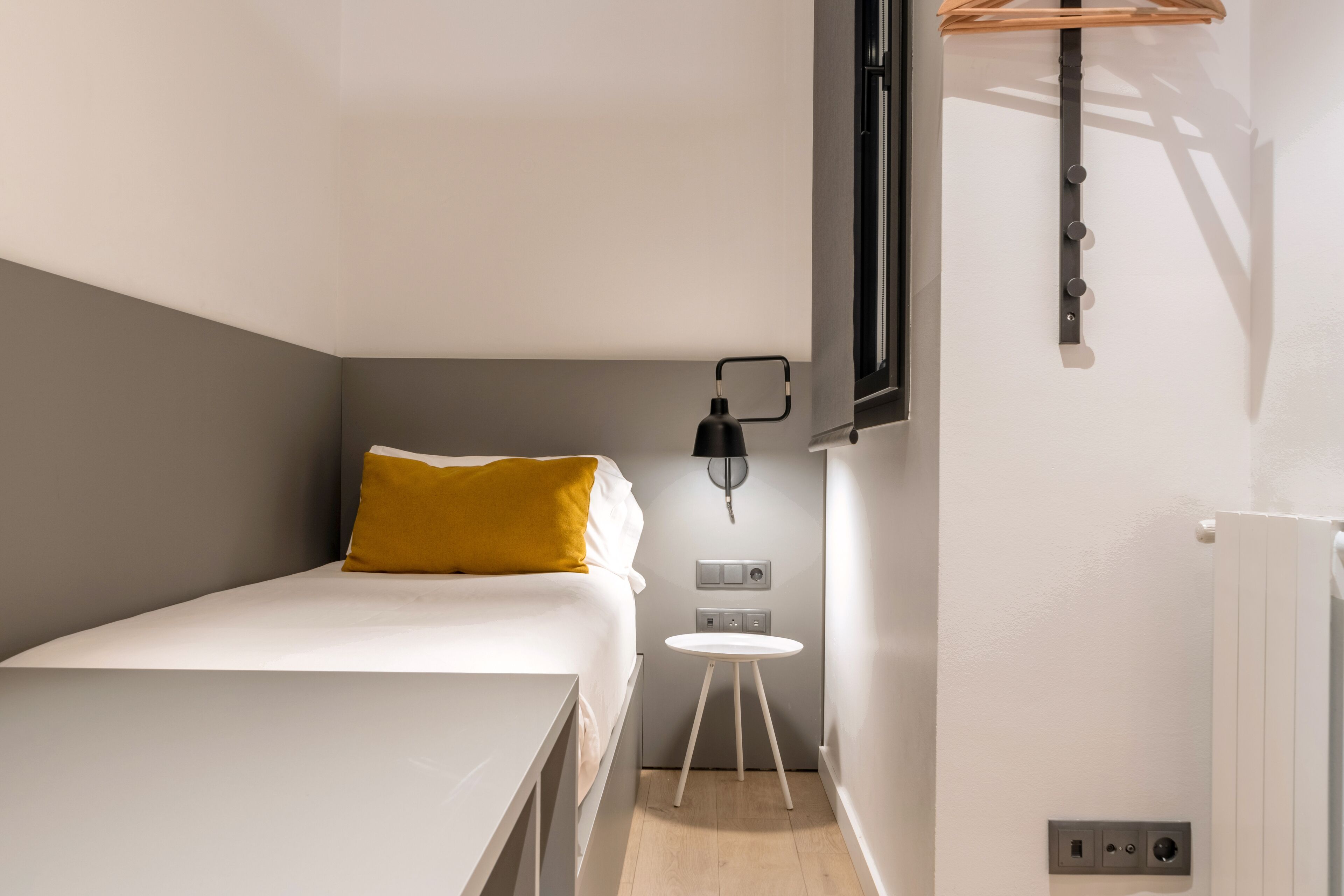 Une petite chambre à coucher moderne, avec un lit simple, une petite table et une lampe fixée au mur.