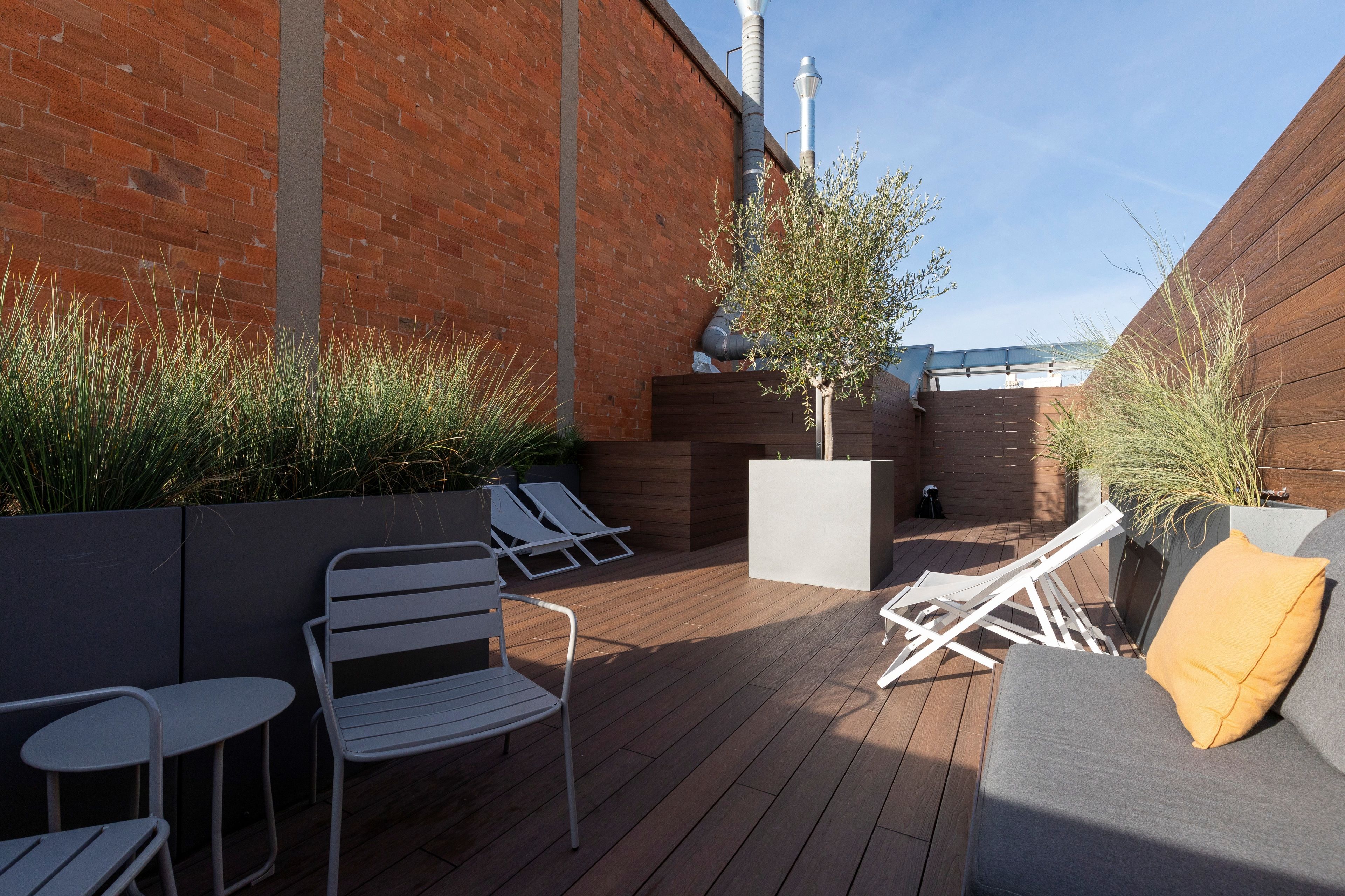 A rooftop patio with a wooden deck, white metal chairs, a gray sofa with a yellow pillow, and a small tree planter.