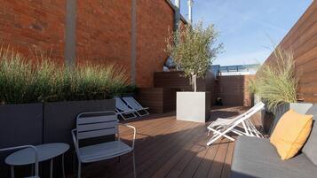 A rooftop patio with a wooden deck, white metal chairs, a gray sofa with a yellow pillow, and a small tree planter.