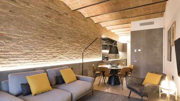 Ein modernes Wohnzimmer mit einem grauen Sofa, gelben Kissen, einem Essbereich mit rundem Tisch und Stühlen sowie einer Ziegelwand.