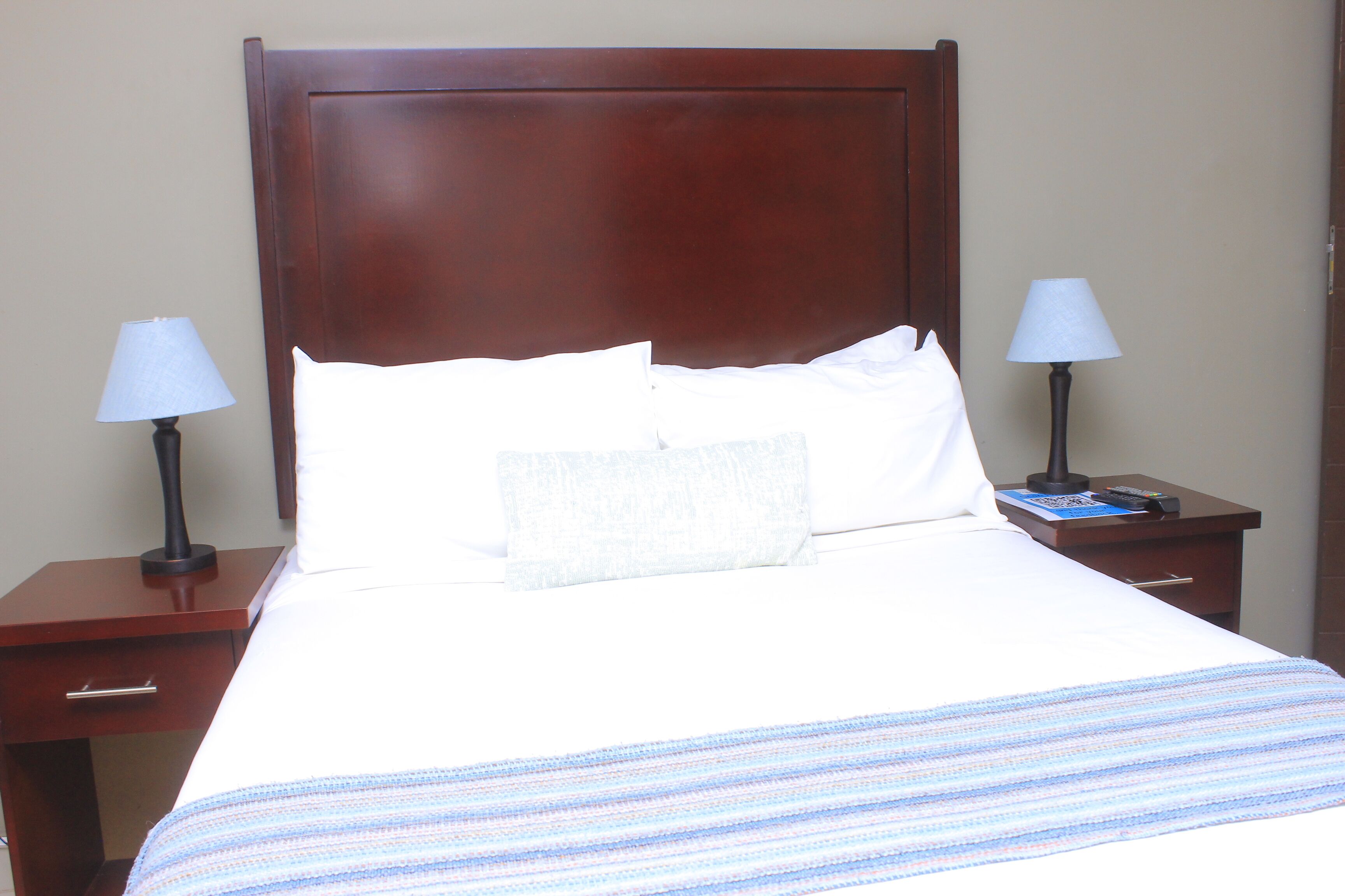 A neatly made bed with white linens and a blue-striped blanket, flanked by two bedside tables with lamps and a small drawer.
