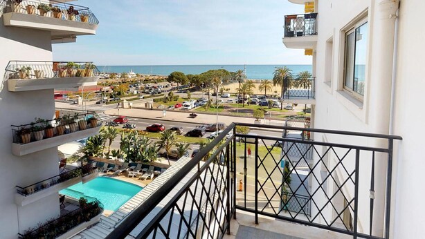 Un hotel con piscina, balcones con plantas y vistas al mar y al estacionamiento.