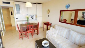 Una sala de estar con un sofá, una mesa de comedor con sillas, una zona de cocina con armarios y una mesa pequeña con un tazón.