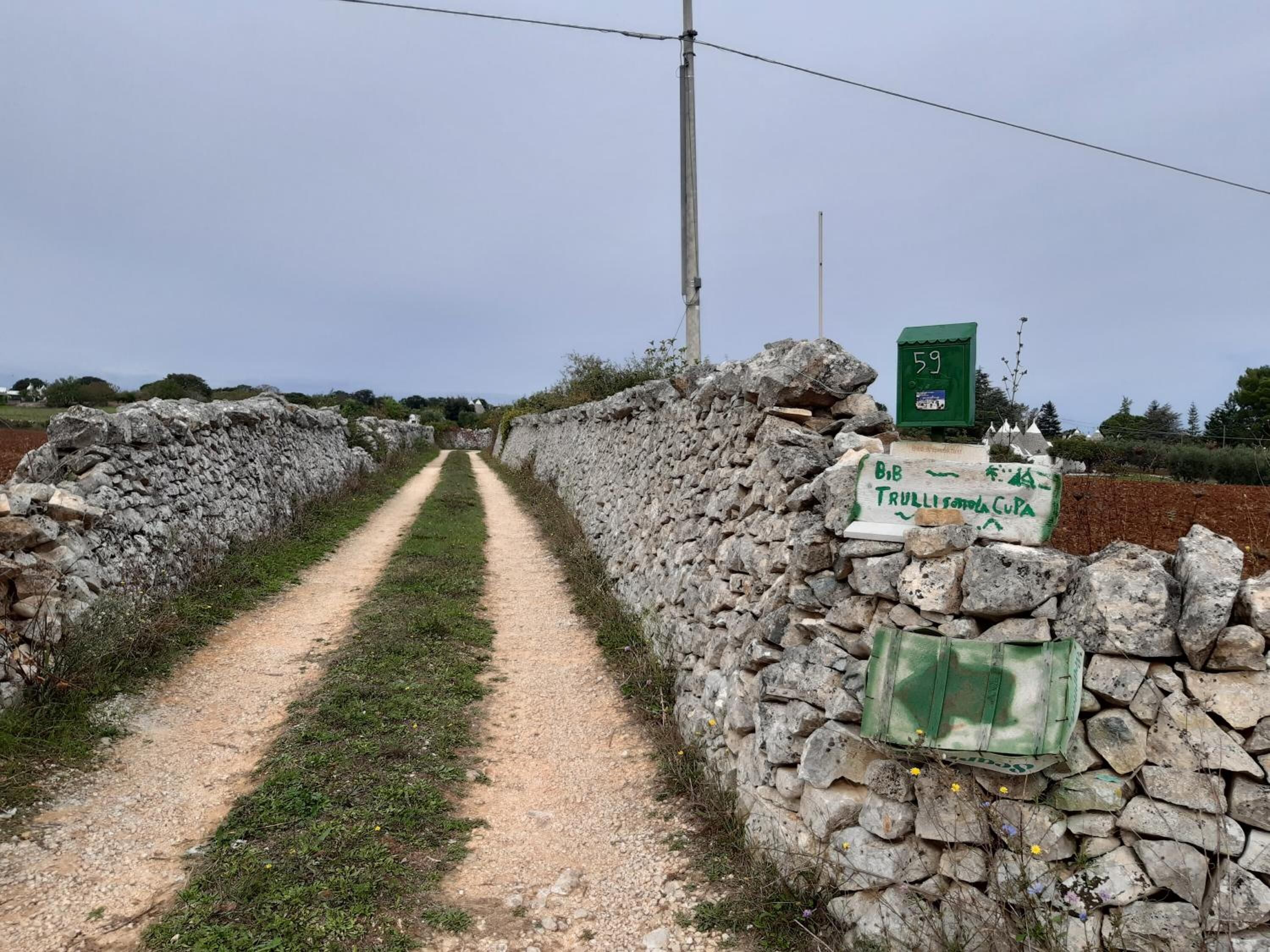A rural dirt path flanked by stone walls, leading to a green signpost with a number 59 and a green box labeled "BIB TRULLIS".