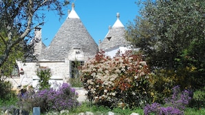 Ein steinernes Haus mit spitz zulaufenden Dächern, umgeben von üppigem Grün und blühenden Pflanzen.