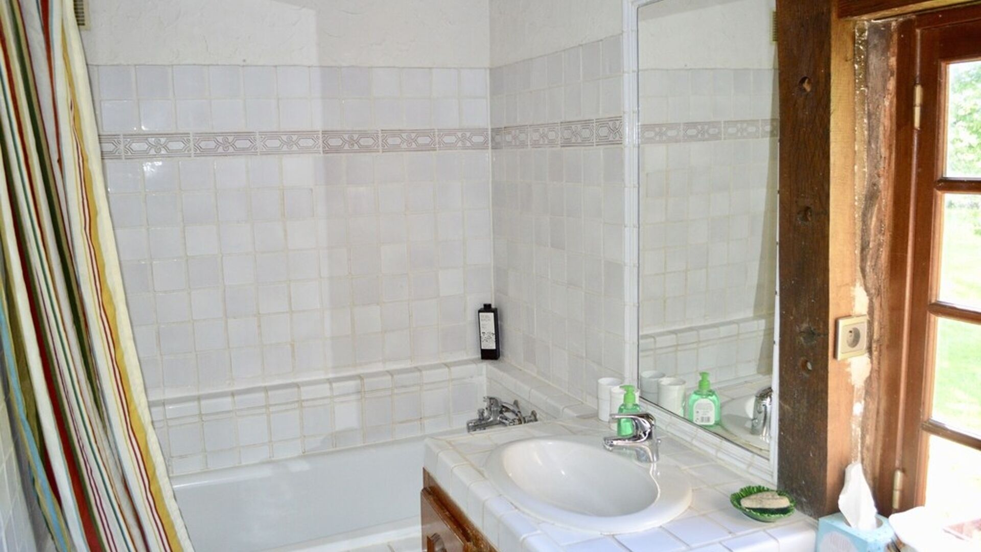 A bathroom with a white sink, mirror, and bathtub. There are toiletries on the counter and a towel hanging on the door.
