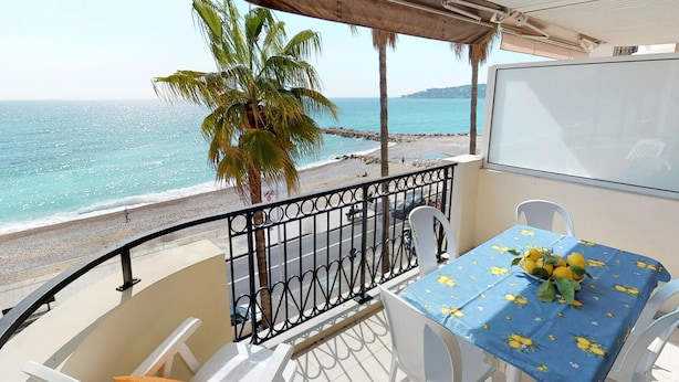 A balcony with a table set for two, overlooking a beach with palm trees and the sea. - Stunning 3BR Apartment with Sea View, Terrace, and Garage in Menton (Menton)