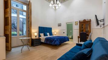 A bedroom with a bed, a blue sofa, a wooden dresser, a small table, a chandelier, and a TV mounted on the wall.