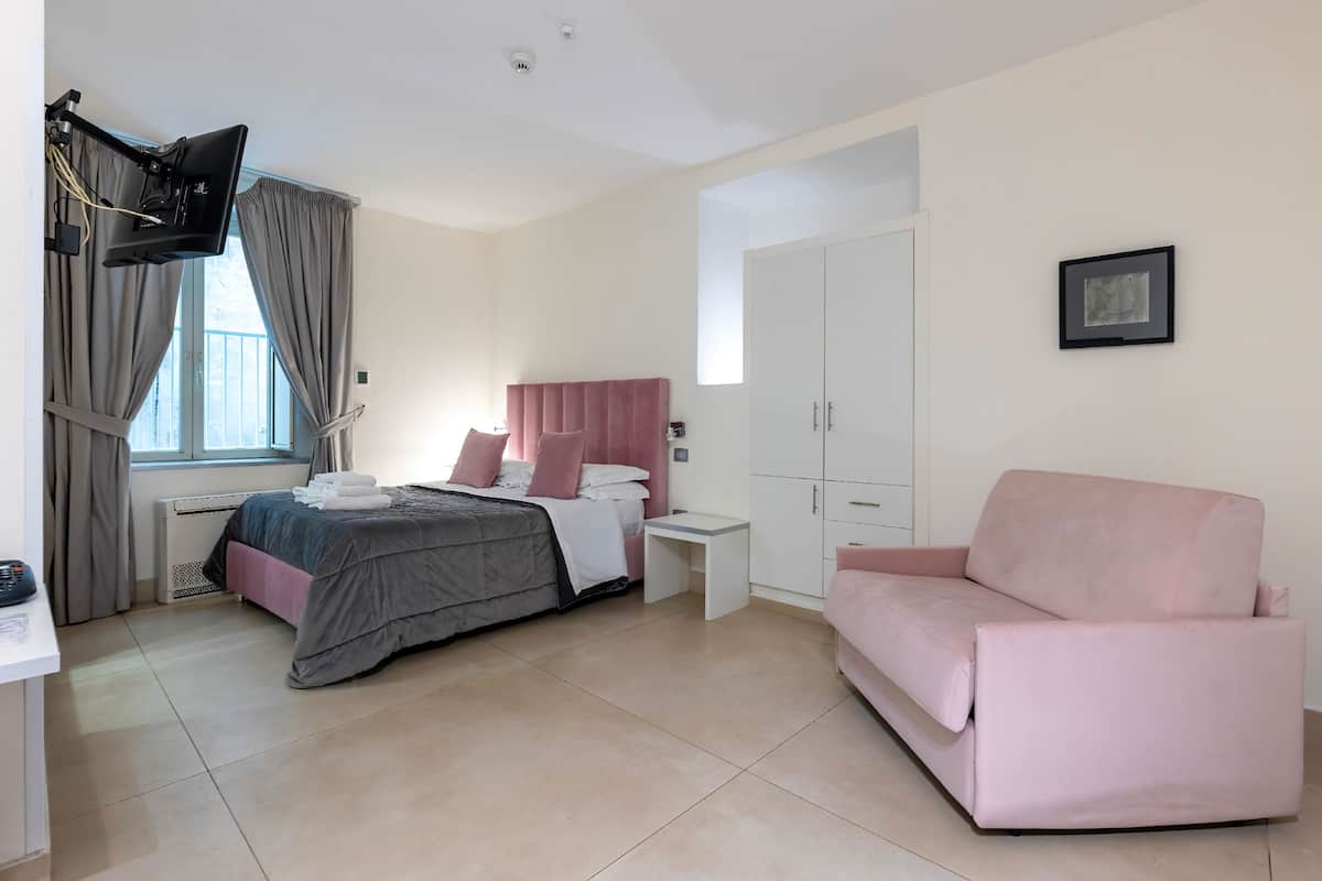 A bedroom with a bed, a desk, a pink sofa, a wardrobe, and a flat-screen TV mounted on the wall.