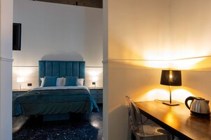 Una habitación de hotel con una cama, lámparas de noche, un escritorio con lámpara y una tetera.
