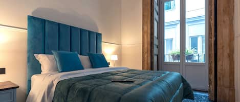 Un lit bien fait, avec une tête de lit bleue et des oreillers, une table de chevet et une fenêtre donnant sur les bâtiments à l’extérieur.