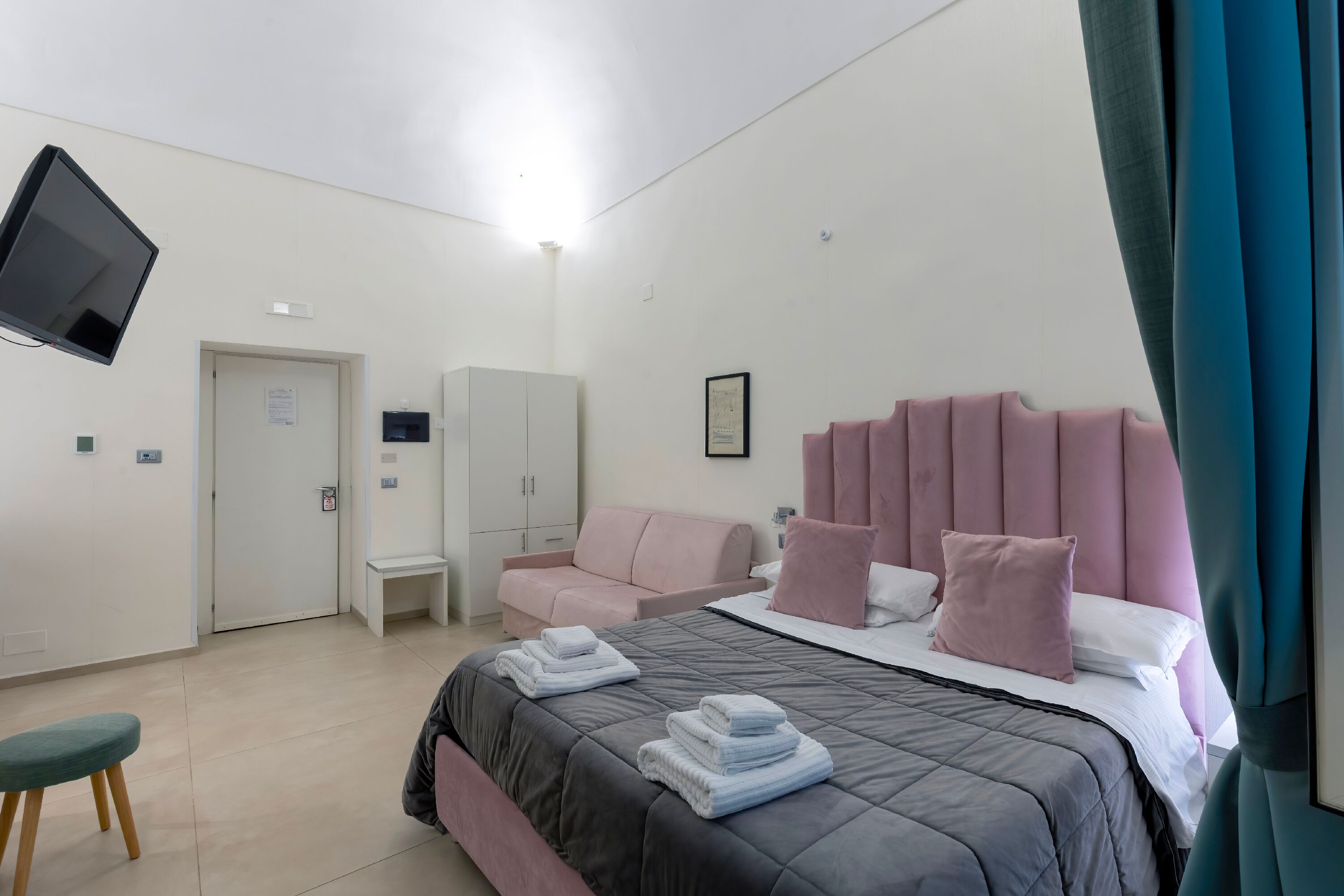 A hotel room with a bed, a TV mounted on the wall, a wardrobe, a bench, and a door.