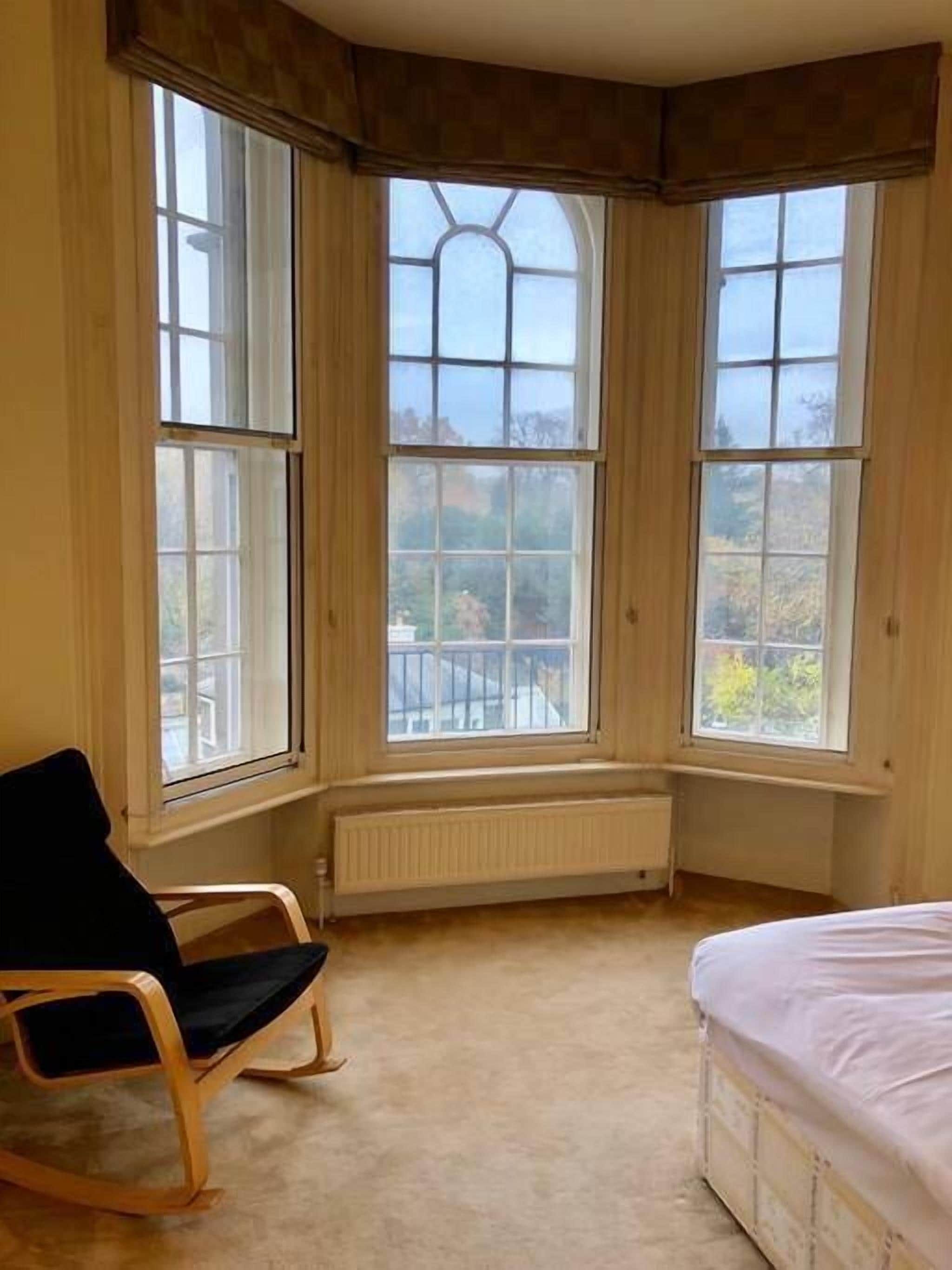 Ein Zimmer mit einem großen Erkerfenster, einem Heizkörper, einem schwarzen Stuhl und einem Bett.