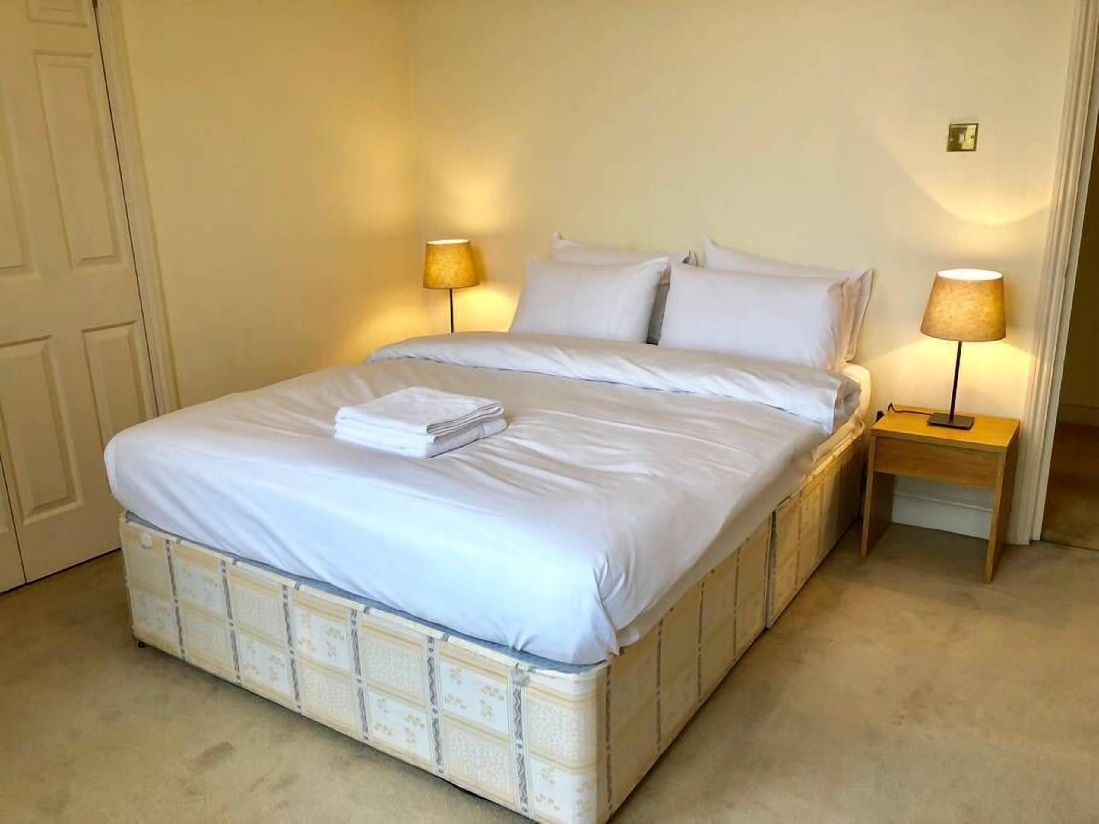 A neatly made bed with white linens and a folded towel, two bedside lamps, and a wooden nightstand.